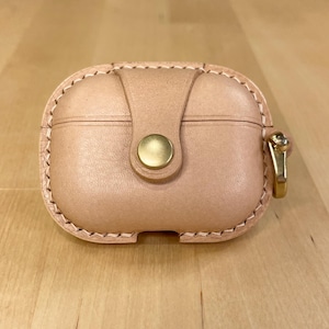Può includere: Una custodia in pelle marrone chiaro con chiusura a bottone dorata e bordo cucito. La custodia è progettata per contenere un piccolo dispositivo elettronico, come un paio di auricolari.