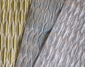 Op de afbeelding: Close-up van drie stoffen stalen met een getextureerd, gewatteerd patroon. De stoffen zijn in tinten goud, grijsblauw en zilver, met een herhalend bladachtig ontwerp. De texturen lijken zacht en pluche, wat suggereert dat het fluweel of een vergelijkbaar materiaal is.