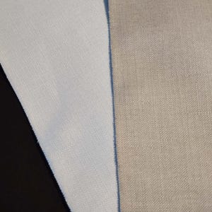 May include: Close-up of two fabric swatches. One is a light blue, the other is a beige color. Both fabrics have a textured weave. The light blue fabric is folded over, revealing a darker blue edge. The beige fabric is flat.