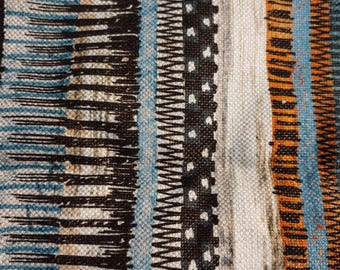 Op de afbeelding: Close-up van een geweven textiel met een verticaal streepjespatroon. Het ontwerp heeft afwisselende banden van zwart, blauw, oranje en wit. De textuur lijkt ruw, met zichtbare draden en een mix van effen en gestreepte lijnen.