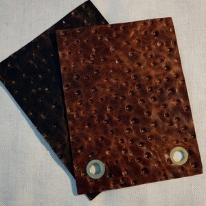 May include: Two pieces of textured leather, one dark brown and one medium brown, are displayed. The leather has a pattern of small, raised bumps. Each piece has a metal grommet in one corner. The leather pieces are overlapping.