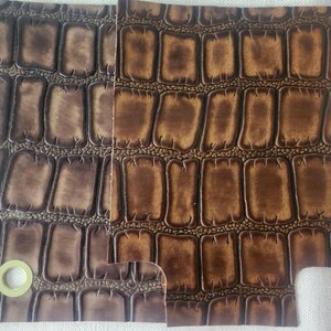 May include: Two rectangular swatches of embossed leather featuring a raised crocodile skin pattern. The left swatch is a dark brown, while the right swatch is a lighter, warm tan brown. A small, circular, light-colored metal grommet is visible.