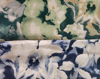 Op de afbeelding: Twee stukken stof met grootschalige bloemenpatronen. Het bovenste gedeelte toont tinten donkergroen, lichtgroen en beige. Het onderste gedeelte heeft een contrasterend ontwerp met donkerblauwe, lichtblauwe en witte bloemmotieven.