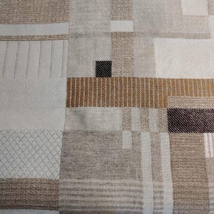 May include: A close-up of a textile with a geometric pattern. The design features squares and rectangles in shades of beige, brown, and white. Some sections have textured details, including stripes and diamond patterns. A small black square is also visible.