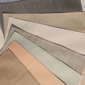 May include: A collection of fabric swatches in various neutral colors, including blue, gray, beige, and peach. The samples are arranged in a fan shape, showcasing the different textures and shades of the material. The fabrics appear to be linen or a similar woven textile.