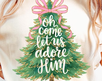 May include: Cream-colored t-shirt featuring a Christmas tree design with the text "Oh come let us adore Him." The tree is green with gold glitter accents and a pink bow at the top. The text is in a cursive font.