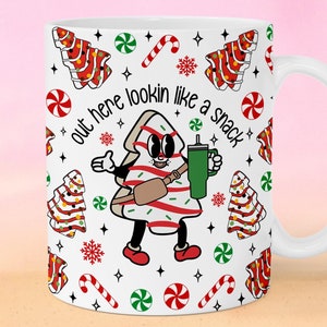 Puede incluir: Taza de cerámica blanca con un patrón repetido de galletas de árbol de Navidad, bastones de caramelo y copos de nieve. La taza presenta un personaje de árbol de Navidad de dibujos animados con el texto "out here lookin like a snack".