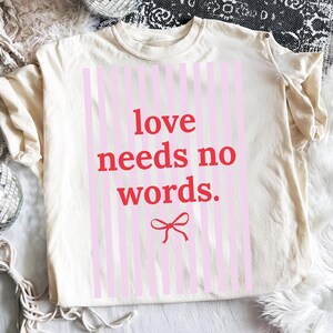 Op de afbeelding: Crèmekleurig T-shirt met de rode tekst "love needs no words" op een roze gestreepte achtergrond. Een rode strik-afbeelding staat onder de tekst. Het shirt ligt op een witte, zachte ondergrond.