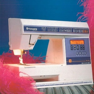 Peut inclure: Une machine à coudre Husqvarna 350 Computer blanche avec un écran numérique bleu et une variété d'options de points. La machine est entourée de plumes roses.