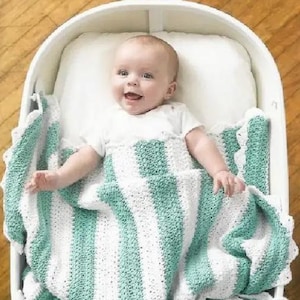 Könnte beinhalten: Ein Baby in einer weißen Babywanne mit einem weißen Kissen, das einen weißen Strampler trägt und mit einer handgefertigten Häkeldecke bedeckt ist. Die Decke hat abwechselnde Streifen in Weiß und Helltürkis.
