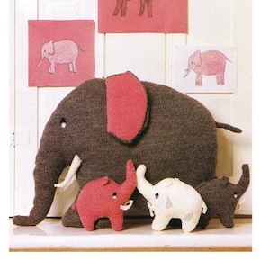 Pode incluir: Um elefante de malha castanho com orelhas cor-de-rosa e um elefante de malha cor-de-rosa ao lado. Há quatro desenhos de elefantes em papel cor-de-rosa na parede atrás dos elefantes de malha.