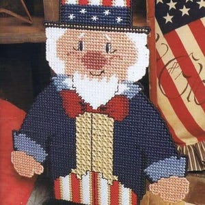 May include: A handmade Uncle Sam figure crafted from plastic canvas. The figure features a top hat with red and white stripes and blue stars, a blue coat, and red and white striped pants. The face is tan with a white beard and a red bow tie. The figure is a patriotic decoration.