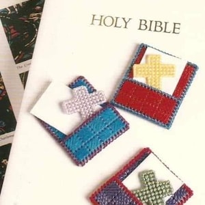 May include: Three small cross-shaped needlepoint ornaments in different colors. The ornaments are in the shape of a pocket with a cross inside. The text "Cross In My Pocket" is at the top of the image. The text "HOLY BIBLE" is on a white book.