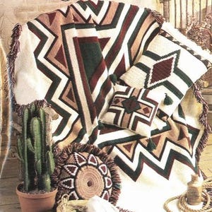 May include: A crocheted blanket with a geometric pattern in shades of brown, red, white, and green. The blanket is folded and draped over a wooden chair. There are other crocheted items in the image, including a pillow and a small rug. The image also includes a cactus in a pot, a skull, and a rope.