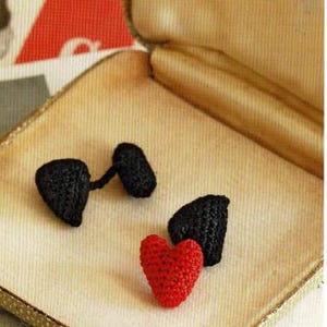 May include: Three crocheted hearts in a small, gold-colored box. Two of the hearts are black and one is red.