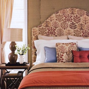 May include: A bedroom with a bed featuring a patterned headboard, a brown blanket, and a red throw. The bed is made with white sheets and pillows, and there are blue and red accent pillows. A brown end table with a lamp and flowers sits next to the bed.