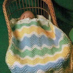 Könnte beinhalten: Ein Baby schläft in einem Weidenkorb, bedeckt mit einer gehäkelten Babydecke mit wellenförmigen Streifen in Blau, Grün, Gelb und Weiß.