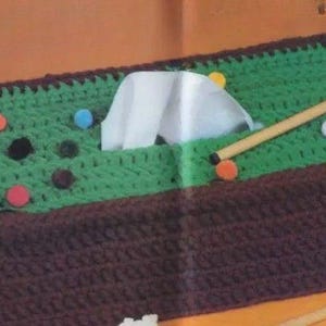 Puede incluir: Una funda para caja de pañuelos de tejido de punto verde y marrón con forma de mesa de billar con un taco y bolas.