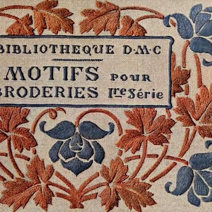 May include: A vintage book cover with the title "BIBLIOTHEQUE D.M.C MOTIFS POUR BRODERIES 1re SERIE" in blue text on a white background. The cover features a floral design with orange and blue embroidered flowers.