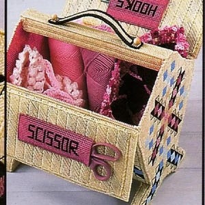 May include: A beige plastic storage box with a pink and black geometric pattern on the side. The box has a handle and is open to reveal pink yarn, lace, and a pink scissor holder with the word "SCISSOR" embroidered on it. The box also has a pink label with the word "WOOL" embroidered on it.