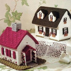 May include: Two handmade miniature houses. One has a red roof and the other has a brown roof. Both houses are white with decorative windows and doors. The houses are sitting on a decorative surface.