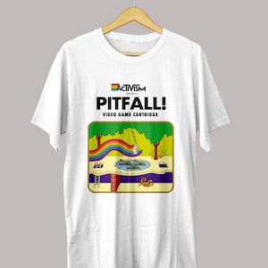 May include: White t-shirt featuring a colorful graphic from the video game Pitfall! The design includes a rainbow, trees, a ladder, and a crocodile-filled pond. The text "ACTIVISM PRESENTS PITFALL! VIDEO GAME CARTRIDGE" is printed above the graphic.
