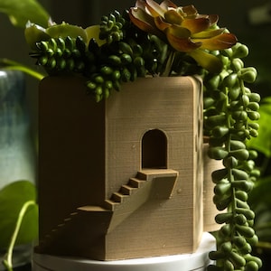 Puede incluir: Una maceta de color marrón claro, de estilo arquitectónico, con una escalera en miniatura y una puerta arqueada. La maceta está llena de una variedad de suculentas verdes y marrones, incluyendo hileras de perlas. La maceta se encuentra sobre una base redonda blanca.