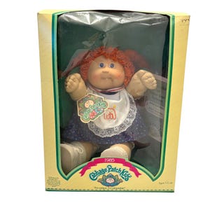 May include: A 1985 Cabbage Patch Kids doll with red yarn hair and blue eyes, wearing a white bib with a red elephant graphic and a navy blue dress with white polka dots. The doll is packaged in a rectangular cardboard box with yellow borders.