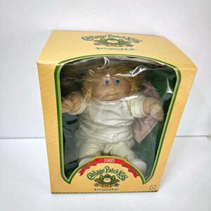 May include: A Cabbage Patch Kids doll with blonde yarn hair and blue eyes, wearing a plain white outfit. The doll is encased in a yellow cardboard box with a clear plastic window, featuring the text Cabbage Patch Kids and the year 1985.