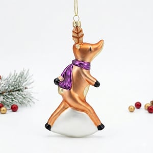 Puede incluir: Adorno navideño decorativo con forma de reno. El reno es de color cobre metálico, con una bufanda morada y pezuñas negras. El adorno está suspendido por un hilo dorado y tiene una base blanca.