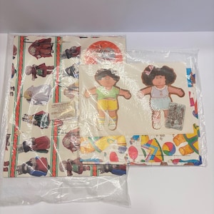 May include: A Cabbage Patch Kids gift wrap set in clear plastic packaging. The set includes a large sheet of wrapping paper featuring repeating illustrations of dolls, and a sheet of two die-cut paper dolls with a green shirt and yellow shorts.