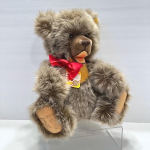 May include: A plush teddy bear with long, shaggy brown fur, a dark brown snout, and a bright red ribbon tied around its neck. A small yellow and white tag with red text and a logo is attached to the ribbon. The bear has light tan paw pads.