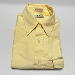 Puede incluir: Camisa abotonada amarilla pálida doblada con cuello abotonado. La camisa tiene un bolsillo en el pecho y lleva el logotipo "L.L.Bean". La camisa está hecha de una tela de color claro.
