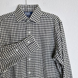 Könnte beinhalten: Ein schwarz-weiß kariertes Button-Down-Hemd von Polo by Ralph Lauren. Das langärmlige Hemd hängt an einem Kleiderbügel. Das Hemd hat einen klassischen Kragen und geknöpfte Manschetten.