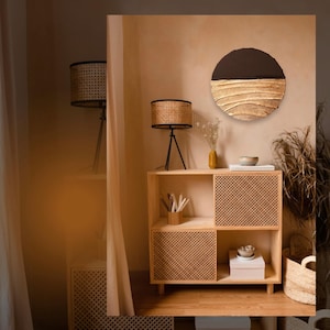 May include: A round wall art piece with a dark brown top and textured gold-colored bottom. The art is displayed above a light wood cabinet with woven door panels. A woven lamp and other decorative items are also visible.