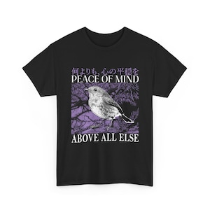 Op de afbeelding: Een zwart t-shirt met een grafische print van een vogel op een tak tegen een paarse achtergrond. De tekst "何よりも、心の平穏を", "PEACE OF MIND" en "ABOVE ALL ELSE" staat in witte en paarse letters boven en onder de vogelillustratie.
