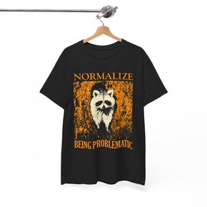 Puede incluir: Camiseta negra con un gráfico de mapache y el texto "NORMALIZE BEING PROBLEMATIC" en naranja. El mapache es blanco y negro, sobre un fondo naranja texturizado. La camiseta está colgada en una percha de madera.