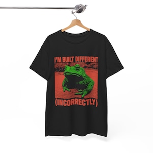 Può includere: Maglietta nera con una grafica rossa e verde di una rana. Il testo "I'm built different (incorrectly)" è stampato sulla maglietta.