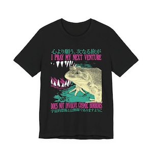May include: A black short-sleeved t-shirt featuring a graphic of a frog and a set of sharp teeth. The text reads I PRAY MY NEXT VENTURE DOES NOT INVOLVE COSMIC HORRORS, accompanied by Japanese characters in teal and pink ink on the front of the garment.