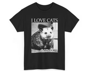 May include: Black t-shirt featuring a grayscale image of an opossum and the text "I LOVE CATS" in a white, bold font. The opossum is centered and appears to be sitting on a wooden surface.