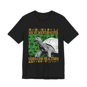 May include: A black short-sleeved t-shirt featuring a graphic of a tortoise against a green background with multiple eyes. The text reads "MAY MY DOUBTERS SUFFER" and "VIDEO-LESS MEALTIMES" in yellow, along with Japanese characters above and below.