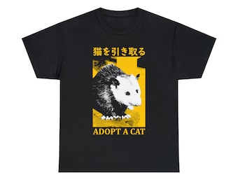 Op de afbeelding: Zwart T-shirt met een gele afbeelding van een opossum en de tekst "猫を 引き取る" in het Japans, wat in het Nederlands "Adopteer een kat" betekent. De tekst "Adopteer een kat" staat ook in het geel onder de opossum.