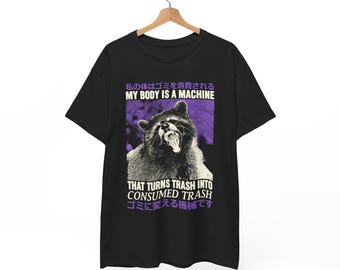 Op de afbeelding: Zwart t-shirt met een wasbeer-afbeelding en de tekst "MY BODY IS A MACHINE THAT TURNS TRASH INTO CONSUMED TRASH" in wit en paars. De wasbeer is afgebeeld met open mond.