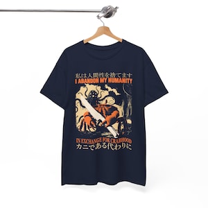Puede incluir: Camiseta azul marino con una ilustración de un cangrejo con una espada. El texto dice "I ABANDON MY HUMANITY" en inglés y japonés. El gráfico es de color naranja y crema.