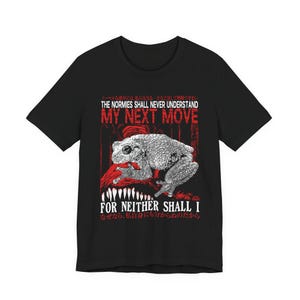 May include: A black short-sleeved t-shirt featuring a white graphic of a frog perched on a red textured shape. The text reads: THE NORMIES SHALL NEVER UNDERSTAND MY NEXT MOVE FOR NEITHER SHALL I, accompanied by Japanese characters above and below.