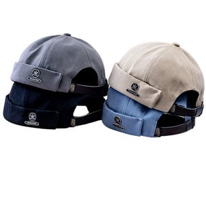 May include: Four docker hats in grey, black, beige, and blue are stacked. Each hat features a short brim and a small emblem. The hats are made of soft fabric and have a strap at the back.