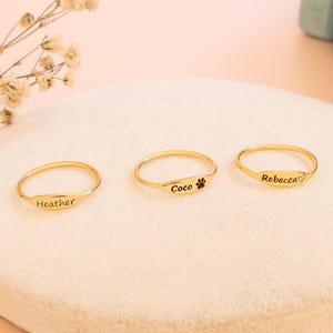 Könnte beinhalten: Drei goldfarbene Ringe mit Gravuren. Einer trägt den Namen "Heather", ein anderer einen Pfotenabdruck und "Coco", der dritte "Rebecca". Sie liegen auf einer weißen, runden Oberfläche vor einem hellrosa Hintergrund.