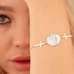 Könnte beinhalten: Ein silbernes Armband mit einem runden Münzenanhänger und zwei Kreuzanhängern. Der Münzenanhänger hat ein detailliertes Design.