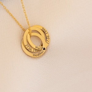 May include: A gold-colored necklace with three interlocking rings. Each ring has engraved text, including names and dates. The necklace is displayed on a white cloth against a blue background.