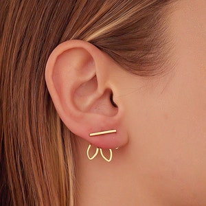 May include: A pair of gold earrings with a simple bar design and a delicate, open-loop design. The earrings are worn in the upper lobe of the ear.